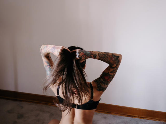 JinaMoon, back to camera, black bra, arms tattooed, long brown hair, hands in hair, sitting on carpet, wooden baseboard, light wall, natural light, sexy, tattoos visible.