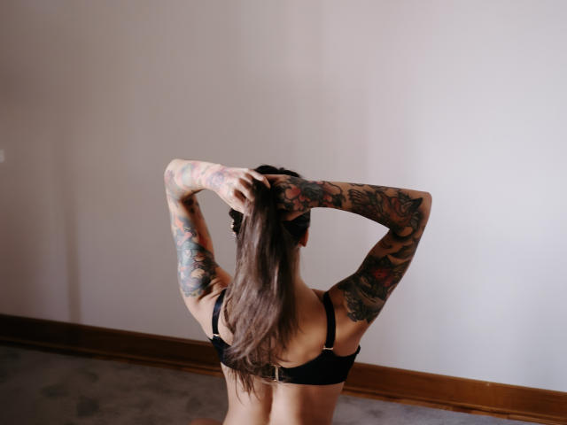 JinaMoon with arms raised, long brown hair, black bra, and full sleeve tattoos, standing against a white wall, wooden floor, and baseboard. Visible back and tattooed arms.
