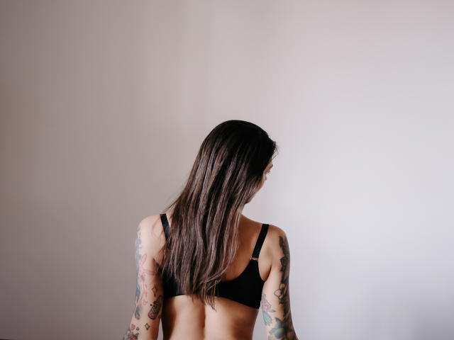 JinaMoon's back facing camera, long brown hair, black bra, tattooed arms, pale skin, standing against a white wall, soft natural light, slim build, small tits.
