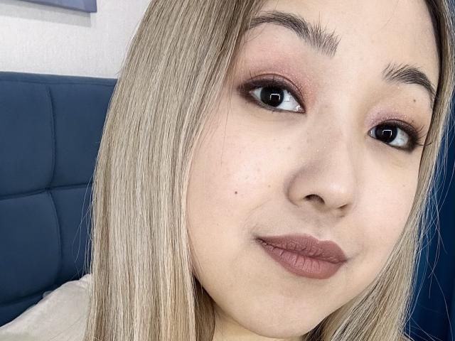 RenyLime has light skin, blonde straight hair, and brown eyes with smoky pink eyeshadow. She's smiling with full pink lips, wearing a beige top, and sitting on a blue chair.