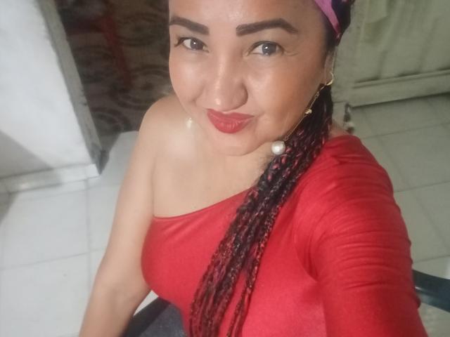 MarieStormy with dark braids, red lipstick, and a pearl earring, wearing a red off-shoulder top, smiling at the camera, showing her bare shoulder and small, cute tits.