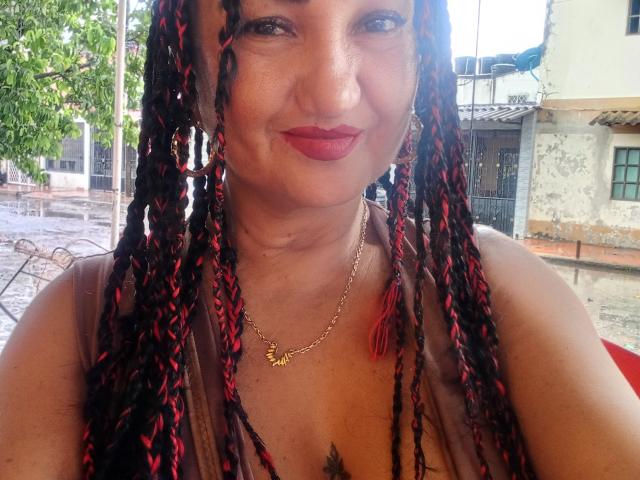 MarieStormy with dark braided hair, red and black highlights, wearing a gold necklace, red lipstick, and a sleeveless top, showing her tits and a small tattoo on her chest.