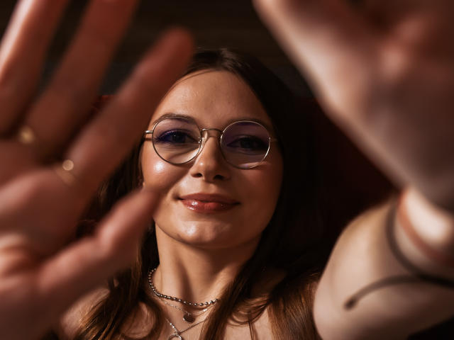 EllyBrune with shiny skin, round glasses, and a necklace, smiling at blurred hands in the foreground. She's got brown hair and a cute face. Her tits are slightly visible.