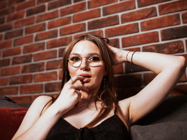 EllyBrune with glasses, finger in mouth, black lace top, hand on head, brick wall, gold bracelets, necklace, red cushion, brown hair, fair skin, sexy, thoughtful expression.