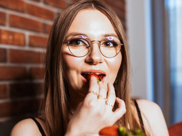 EllyBrune with glasses, biting a strawberry, fingers touching her lips, light brown hair, purple eyeliner, gold rings, brick wall background, smiling, sexy, cute.