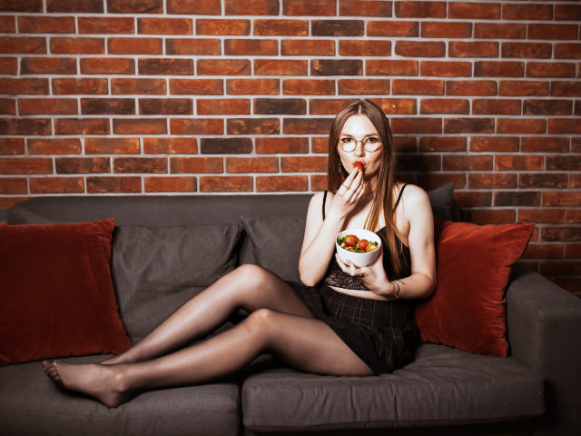 EllyBrune with glasses, eating fruit from a bowl, wearing a black lace top, plaid skirt, and sheer stockings, sitting on a gray sofa with red pillows.