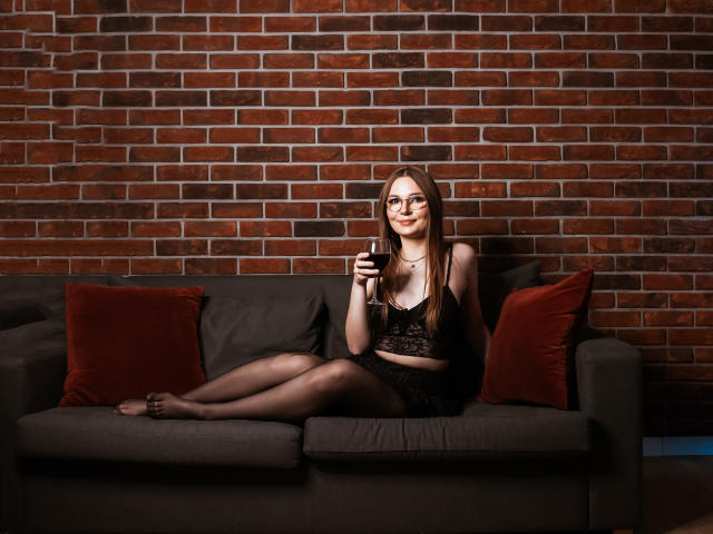 EllyBrune with glasses, black lace bra, and panty, holding wine glass, sitting on gray sofa with red pillows, legs crossed, smiling, brick wall background.