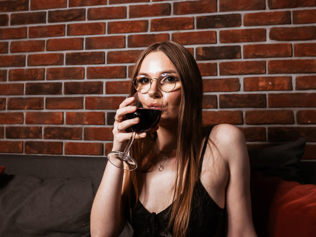 EllyBrune with glasses, sipping red wine, has long brown hair, black lace top, and a necklace, sitting against a brick wall, showing her tits subtly.