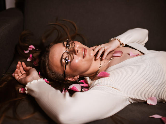 EllyBrune with glasses, lying on dark couch, arms above head, pink petals on chest, white ribbed sweater, soft smile, brown hair, hand near face, relaxed.