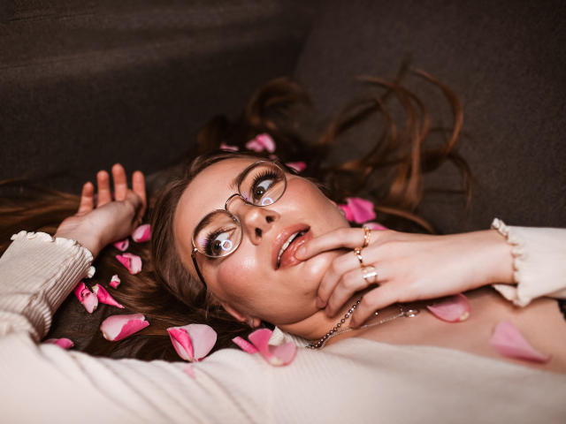 EllyBrune with glasses, lying on a gray couch, pink rose petals in brown hair, fingers touching her mouth, white shirt, gold rings, and a delicate necklace.