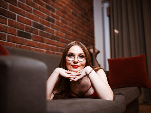 EllyBrune with glasses, red lipstick, and long brown hair, leaning on a gray sofa, hands under chin, red bra, smiling, red and brown pillows, brick wall background.