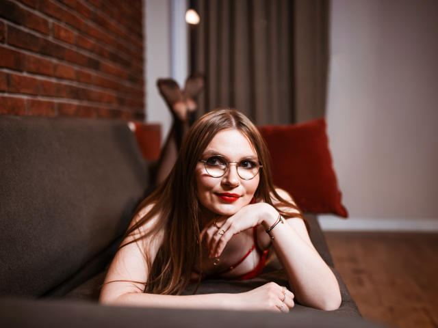 EllyBrune with glasses, red lipstick, and red bra, laying on a gray couch, legs up, showing her ass, smiling, wooden floor, brick wall, brown curtain, red pillow.