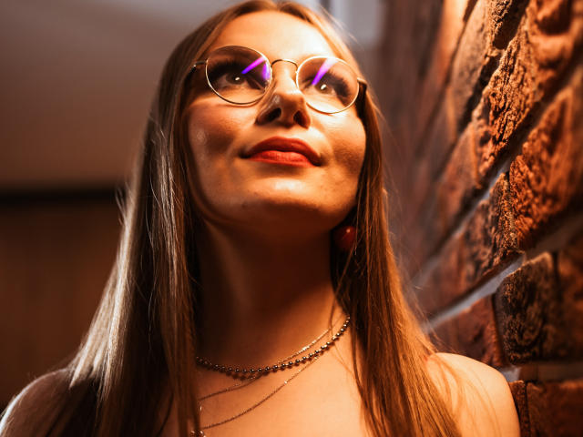 EllyBrune with glasses, looking up, has shiny red lips and a subtle smile, wearing a necklace, against a brick wall, with warm light highlighting her face.