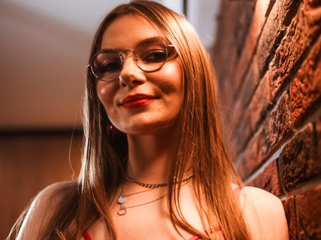 EllyBrune with long blonde hair, red lipstick, glasses, and a necklace, standing against a brick wall, smiling softly, showing shiny skin, wearing a red top.