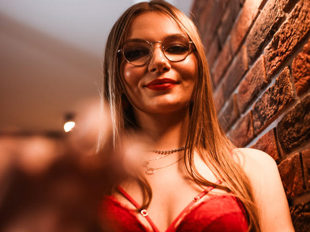 EllyBrune with long blonde hair, red lipstick, and glasses, wearing a red lace bra, smiling at a blurred object in the foreground, against a brick wall.
