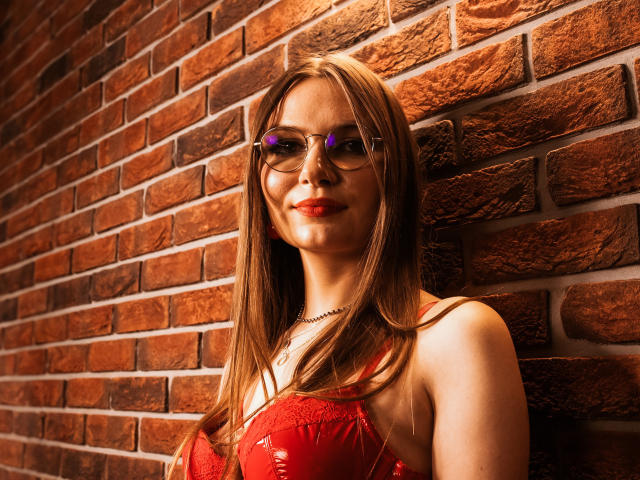 EllyBrune with glasses, red dress, and red lipstick, has her tits slightly visible, standing against a brick wall, smiling, wearing a necklace, brown hair, and purple glasses.