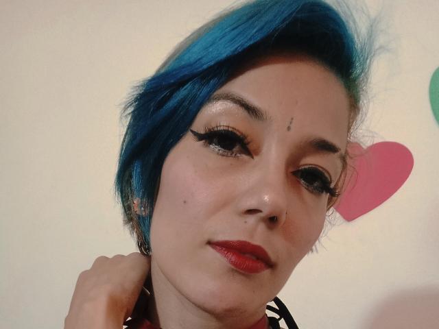 Vero69 with blue hair, black eyeliner, red lipstick, nose piercing, and small beauty mark on her forehead, smiling softly, hand near neck.