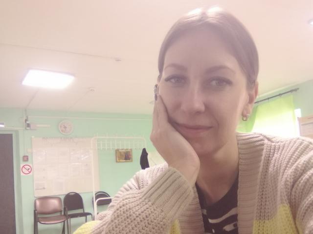 AshleyXHoney with short brown hair, smiling, hand on cheek, wearing a beige and yellow knit cardigan, black shirt with white stripes, in a brightly lit room.
