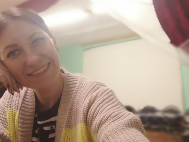 AshleyXHoney with short brown hair, smiling, head resting on hand, wearing a pink and yellow striped cardigan, black shirt with white letters, indoors, bright light in background.