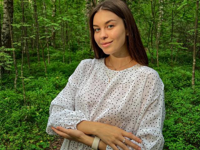 GloryJasmine with long brown hair, wearing a white polka-dot shirt, standing in a forest, arms crossed, smiling softly, green background, natural light, looking cute.