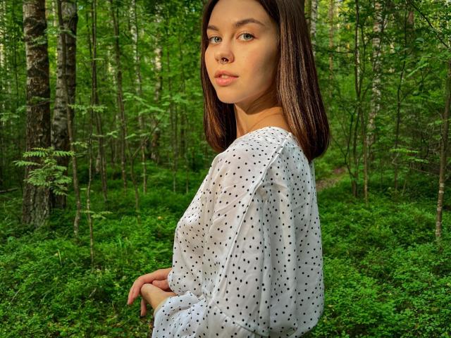 GloryJasmine with shoulder-length brown hair, nose ring, and white polka dot blouse, looking over shoulder in a green forest. Her tits are small, and her ass is not visible.