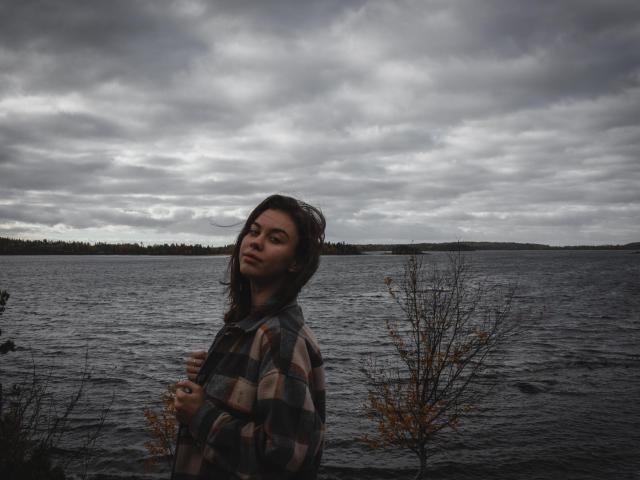 GloryJasmine with long dark hair, wearing a plaid shirt, standing by a dark, cloudy, water-filled scene, looking to the side with a slightly serious, thoughtful expression.