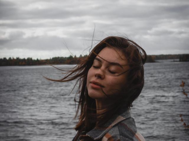 GloryJasmine's face with closed eyes, brown hair blowing, gray plaid shirt, dark water, cloudy sky, and tree line in background, looks relaxed and serene.