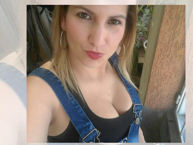 GiuliaAddams with blonde hair, wearing blue denim overalls, black tank top showing her tits, and red lipstick, looking at the camera with small hoop earrings.