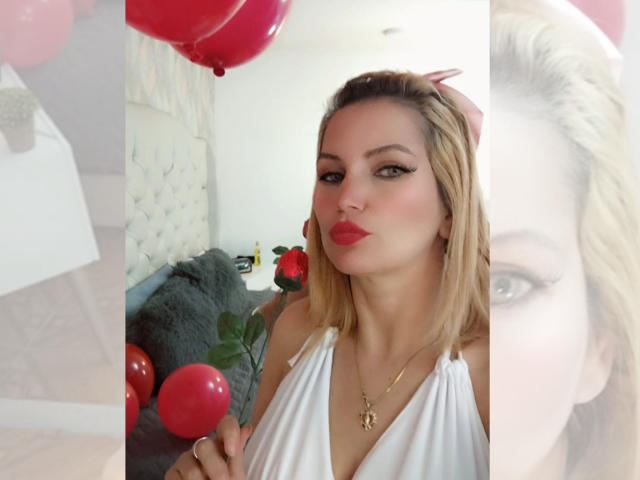GiuliaAddams met blond haar, rode lippenstift, een witte haltertop, een gouden ketting, die rode ballonnen vasthoudt, met een fronsende uitdrukking, grote ogen, subtiele make-up, zichtbare borsten, in een slaapkamer, met een onscherpe overlay over het gezicht..