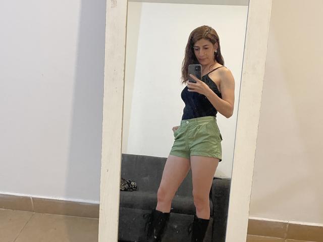 RobertaWood with brown hair, small tits, wearing a black tank top, green shorts, and black boots, taking a mirror selfie, standing in a white-walled room.