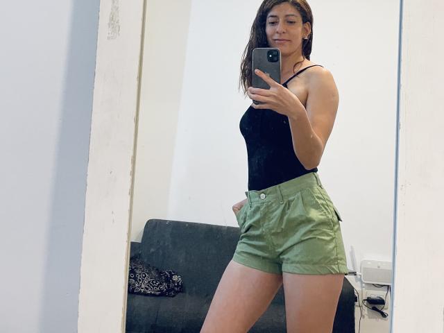RobertWood, with a slim, fit physique, wearing a black tank top and green shorts, taking a mirror selfie, showing her small ass and slim legs.