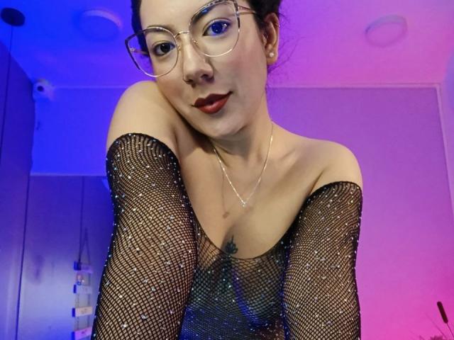 AbbySton mit Brille, trägt ein glitzerndes schwarzes Netzoberteil mit offener Schulter, das ihre kleinen Brüste zeigt und ein Tattoo dazwischen, vor einem violett-blauen Hintergrund..