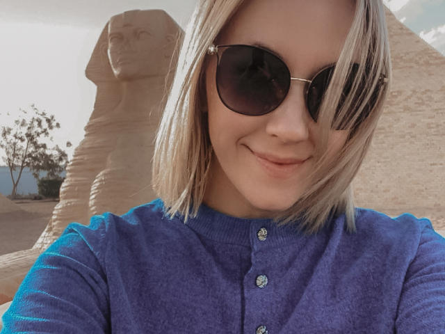 Misstyk with blonde hair, wearing dark sunglasses and a blue button-up shirt, smiling at the camera. Her tits are small, and she's standing in front of a giant stone statue.