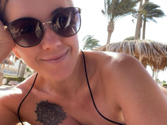 Misstyk con gli occhiali da sole, sorridente, che mostra un tatuaggio sul petto, indossa un top da bikini nero a filo, illuminata dal sole, palme sullo sfondo, capelli castani, pelle chiara..