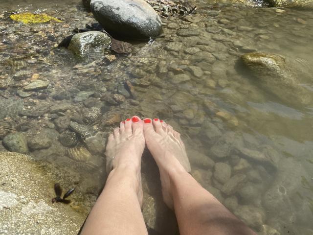 JuliaGarciax's feet with red-painted toenails in clear water, standing on a rocky stream bed, showing her fair-skinned legs and toes.