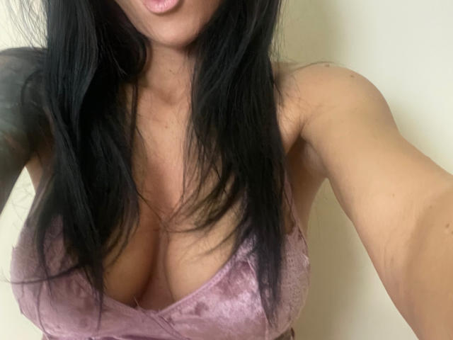 AndraD with long black hair, pink velvet bra, showing her tits, pouty lips, tan skin, and a tattoo on her left arm. Soft lighting, slightly blurred.