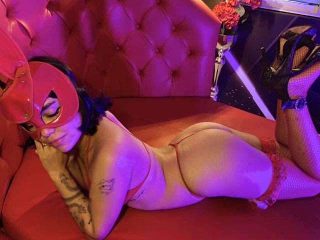 Anni Evans est allongée sur un canapé rouge capitonné, portant un masque rouge, un string rouge, des bas résille et des talons noirs. Ses fesses sont proéminentes, et des tatouages sont visibles sur son bras..