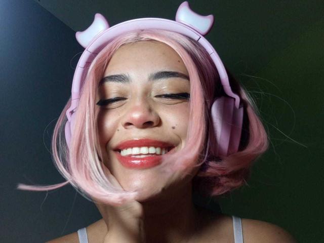 AnniEvans with pink hair and pink headphones, smiling with eyes closed, has a small mole on her nose, wearing a white tank top, and has cute pink devil horns.