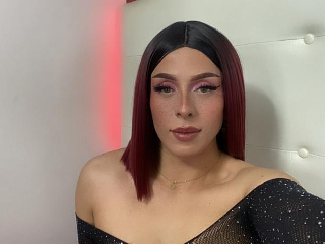 Megan Martell met een donkerrode bob, roze eyeliner en een zwarte top met glitters die van de schouder valt, waardoor haar blote schouders en ketting zichtbaar zijn. Haar borsten zijn gedeeltelijk zichtbaar..