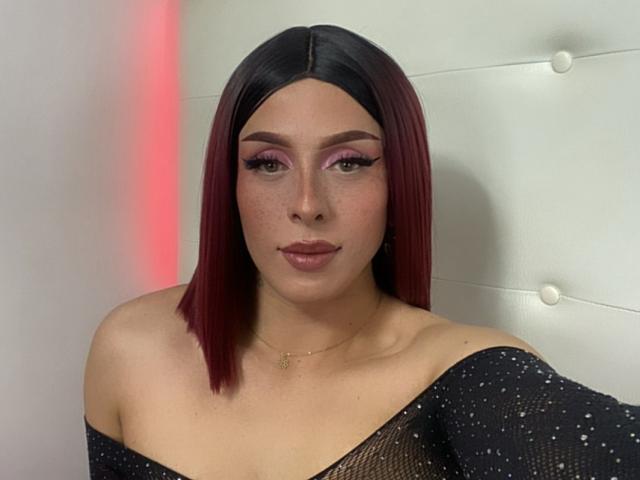 MeganMartell heeft donkerrood, steil bob-haar, roze oogschaduw, lange wimpers, sproeten en draagt een zwart, doorschijnend topje met een open schouder. Haar achterkant is niet zichtbaar. Ze draagt een ketting..