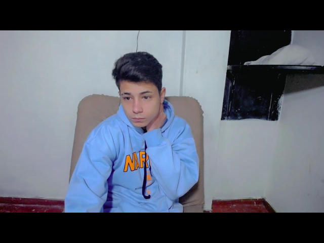 AlaBoyce, young dude with dark hair, wearing a light blue "NIF" hoodie, hand on neck, serious expression, sitting in a beige chair, right wall partially visible.