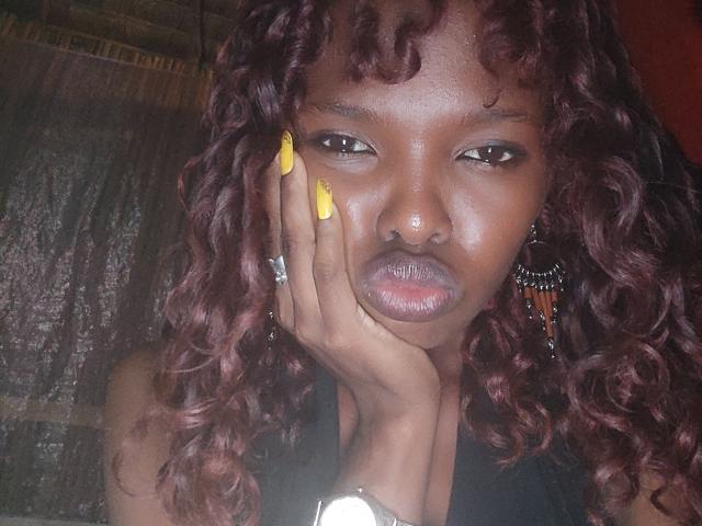 Mureille with dark brown skin, curly red-brown hair, yellow nails, hand on face, nose ring, and black dress, looking serious. She's wearing a ring and a bracelet.