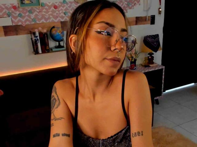 LemoonPeach with glasses, nose ring, and tattoos, wearing a sparkly black top, has a serious face, brown hair, and small tits. She's in a dimly lit room.