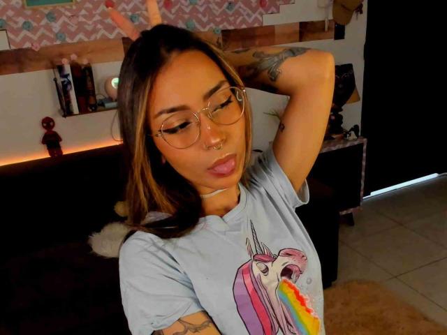 LemoonPeach with glasses, tongue out, wearing a unicorn shirt, arms raised, tattoos visible, nose ring, light skin, brown hair, in a cozy room, looking relaxed.