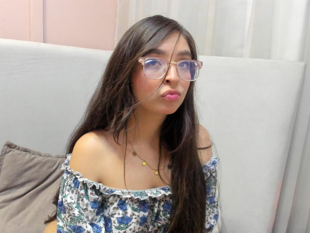 EvaMcAllister, avec ses longs cheveux foncés, son rouge à lèvres rose, ses lunettes et sa robe fleurie à épaules dénudées, faisant légèrement la moue, un collier avec de petites perles jaunes, assise sur un canapé gris..