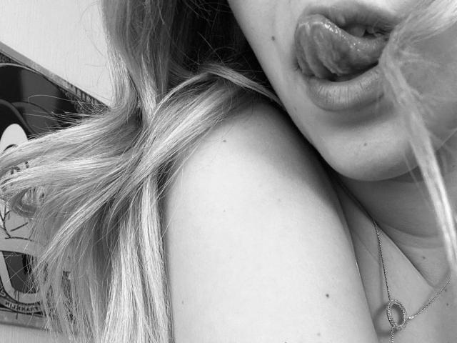 SugarrX's tongue is out, licking her upper lip with shiny, plump lips and a small mole on her cheek. Her blonde hair is partially visible, and she's wearing a necklace.