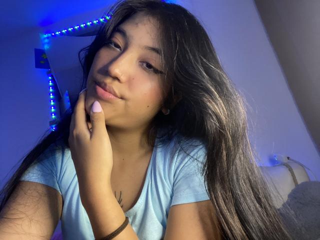 Elizaa met lang zwart haar, een lichtblauwe T-shirt, en roze nagellak, die haar wang aanraakt, lachend, met blauwe LED-stripverlichting op de achtergrond, schattig, mooi, kleine neus, subtiele tatoeage..
