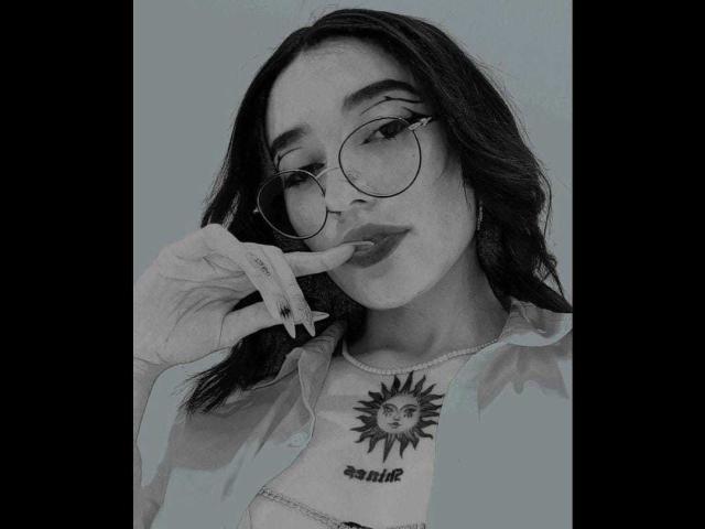 SalomeMille with long dark hair, wearing round glasses and a light shirt, biting her finger, showing a sun tattoo on her chest. Her tits are small, she's cute.