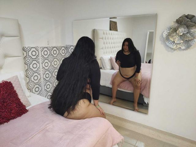 ZoeGiil with long black hair, wearing a black crop top and black thong, sitting on a pink bed, looking at her reflection in a mirror, showing her big ass.