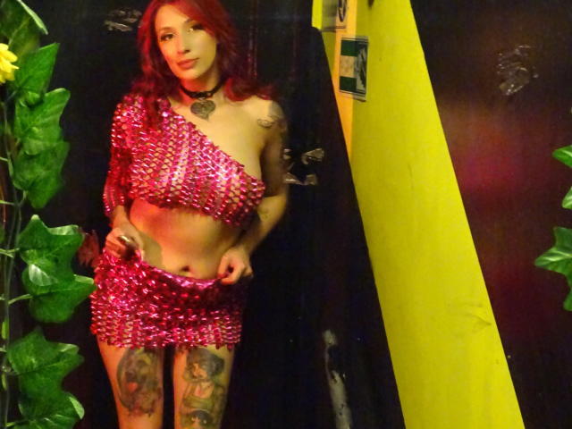 AriadnaFetish69 with red hair, pink sequin one-shoulder mini-dress, showing her tits, tattoos on thighs, choker, and looking at camera, standing next to green plants and yellow wall.